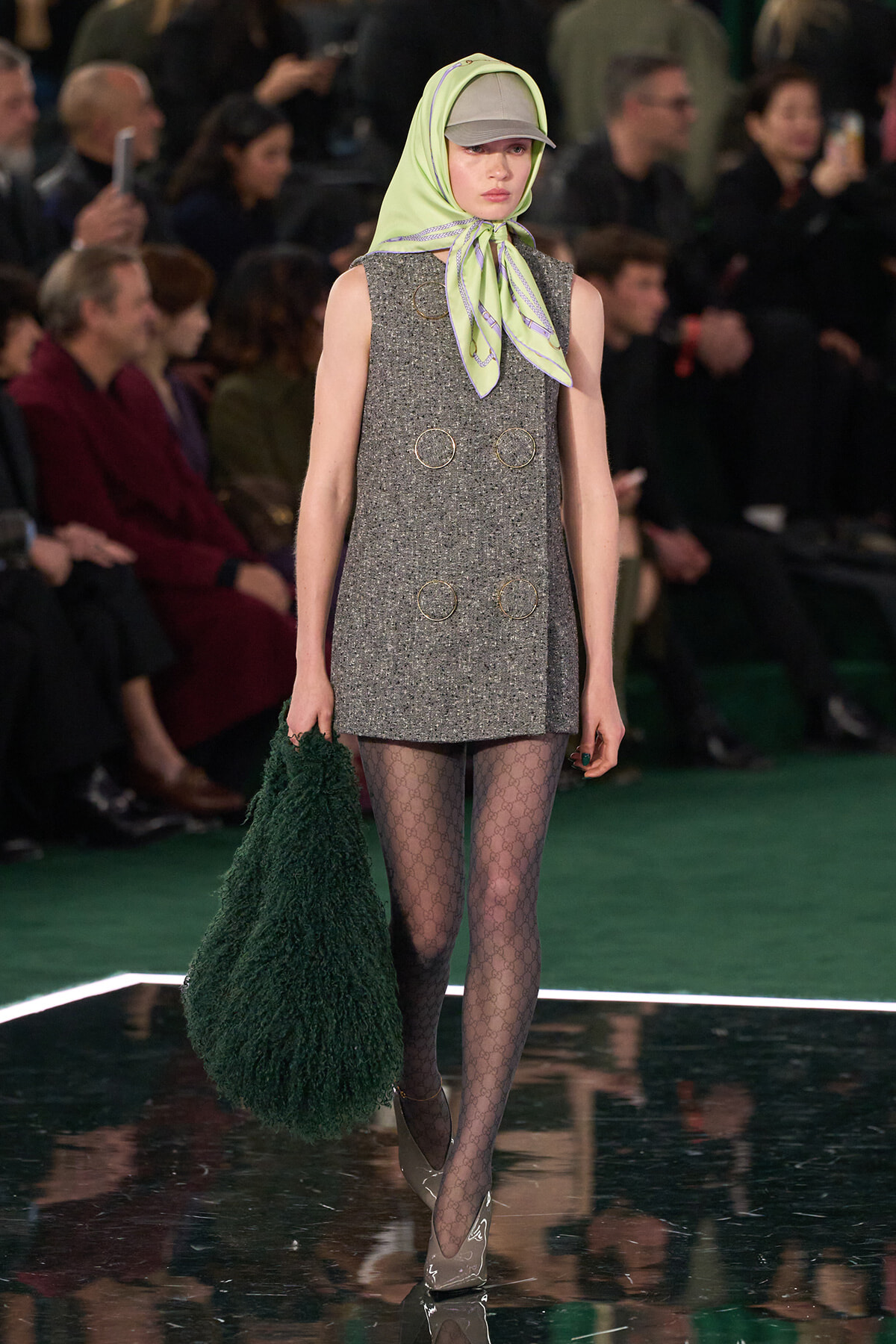 Model walks the runway in a sleeveless gray textured mini dress with circular metal rings, lime scarf tied under a cap, patterned tights, and heels, carrying a dark green fuzzy bag.