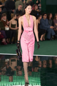 Model walking the runway in a pink satin lace jumpsuit with a plunging neckline, gold chain necklace, black gloves, and a small black handbag.