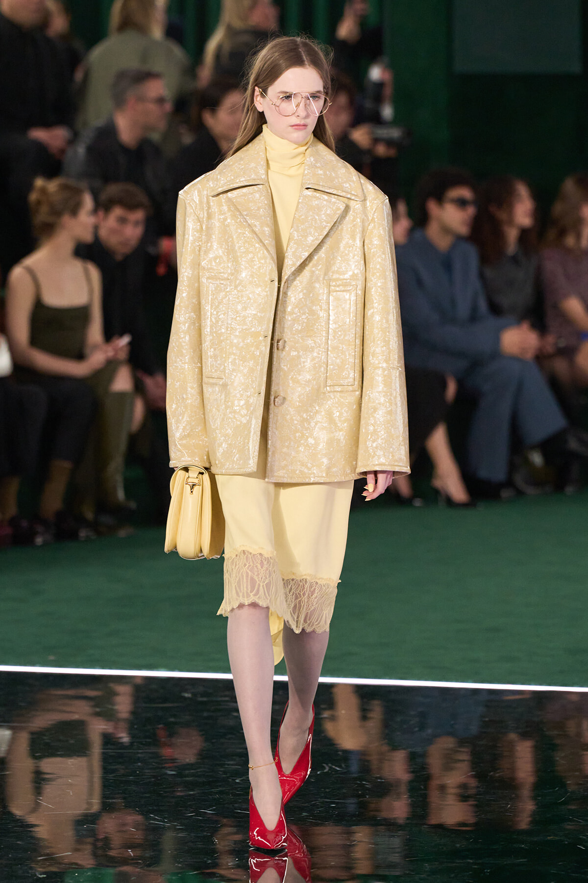 Runway model wearing a pale gold oversized textured coat over a yellow dress with lace hem, carrying a yellow bag and red heels.