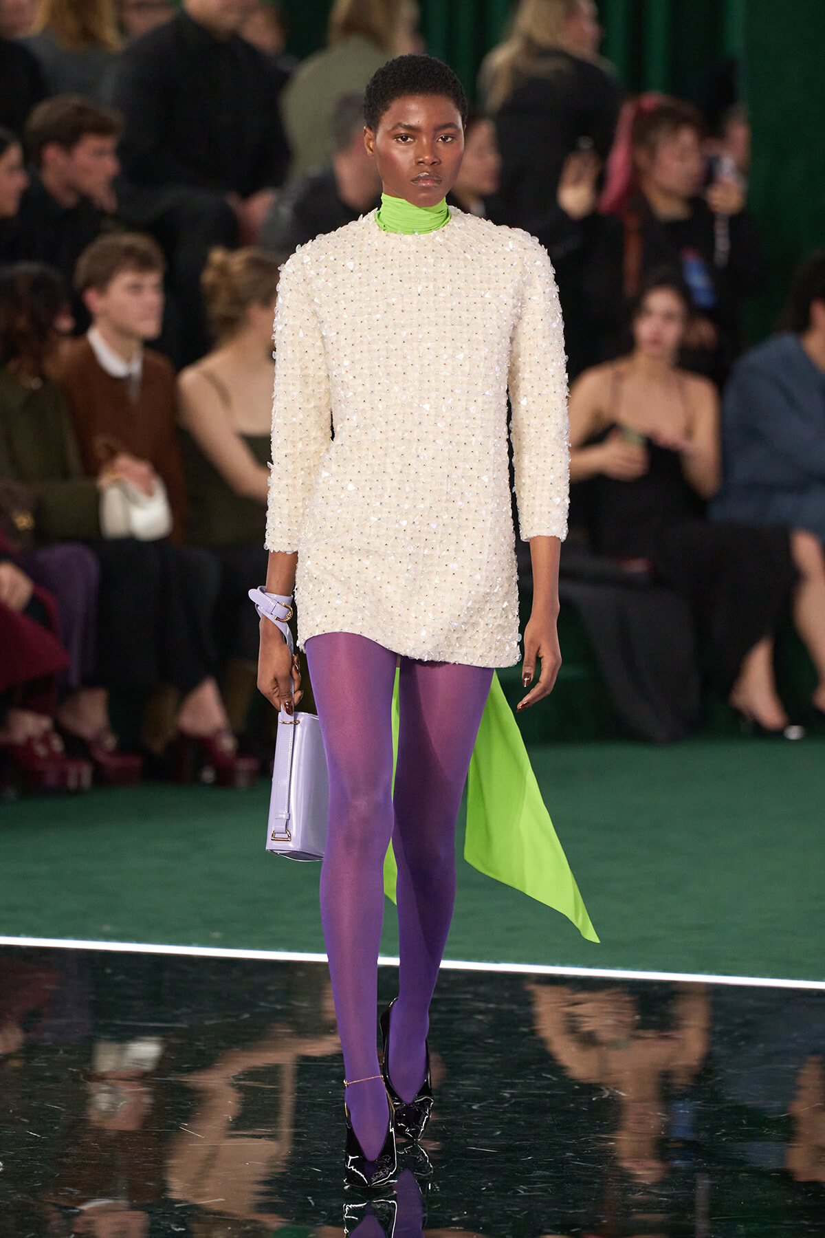 Model on a runway wearing a cream pearl-embellished mini dress with a neon green turtleneck, bright purple tights, and a lavender handbag.