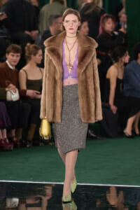 Model walking runway in brown fur coat over lavender lace top and glittery grey pencil skirt with lime green heels and a yellow handbag.