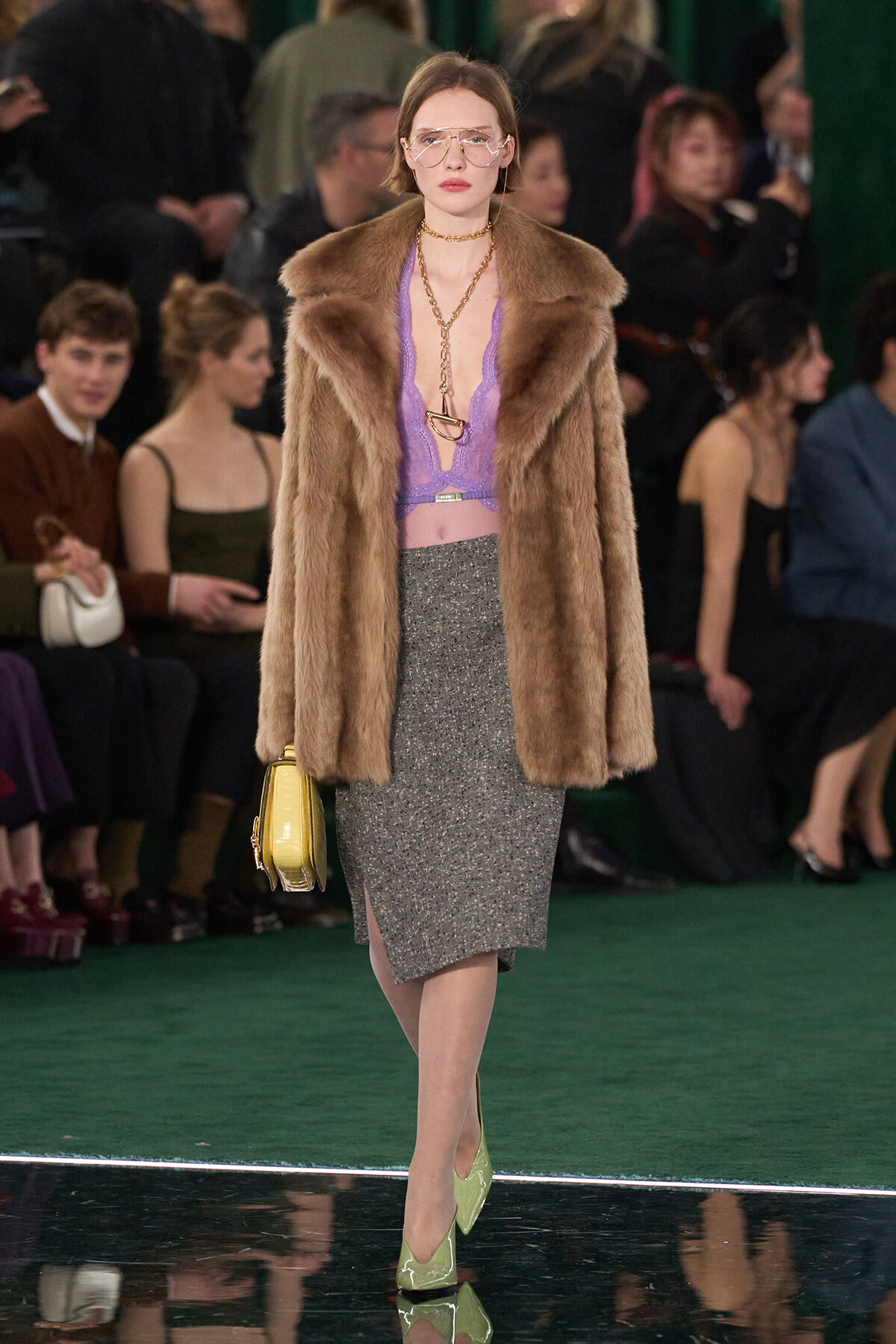 Model walking runway in brown fur coat over lavender lace top and glittery grey pencil skirt with lime green heels and a yellow handbag.