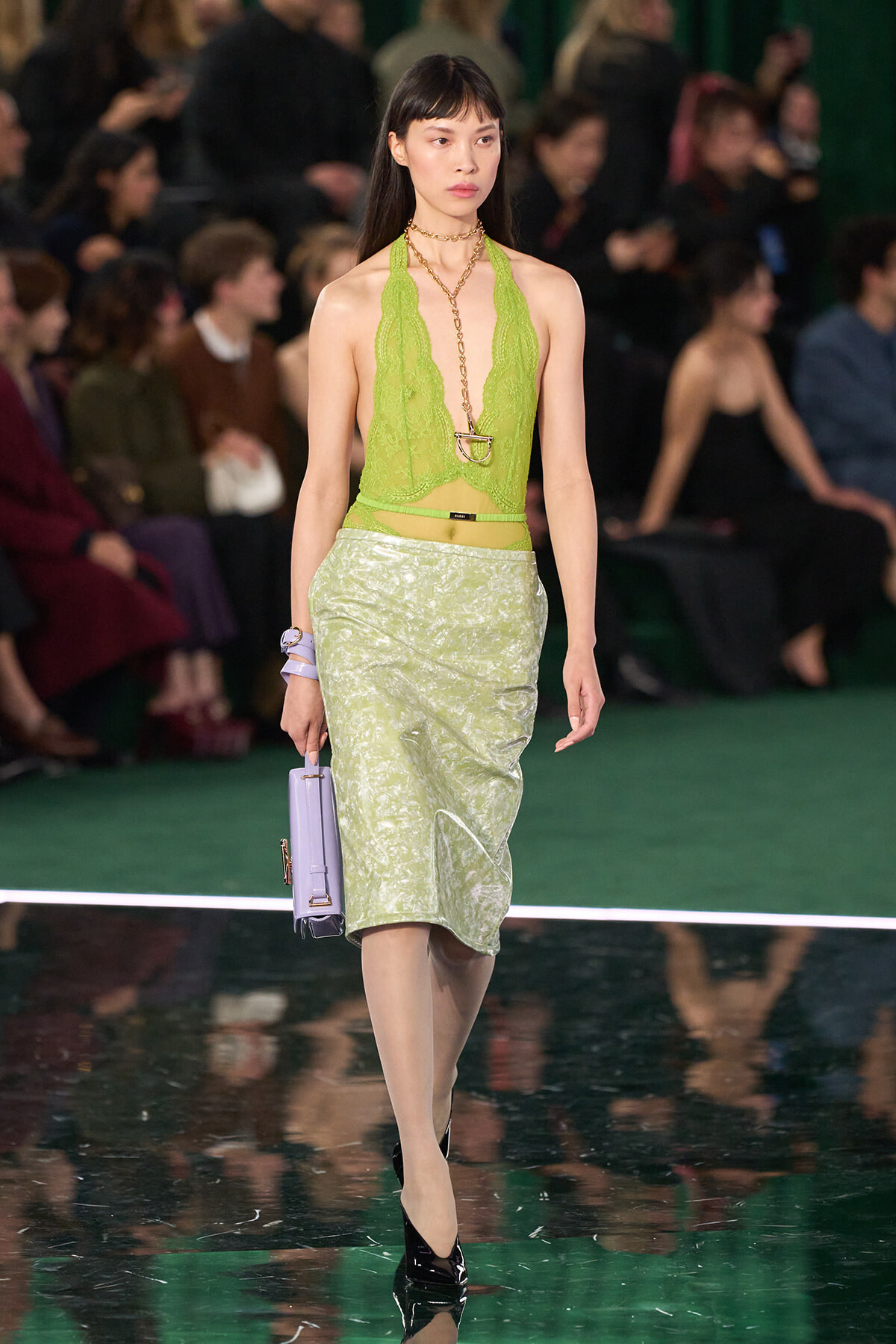 Model walking a fashion show in a lime green lace halter top and pale green knee-length skirt, holding a lavender handbag.