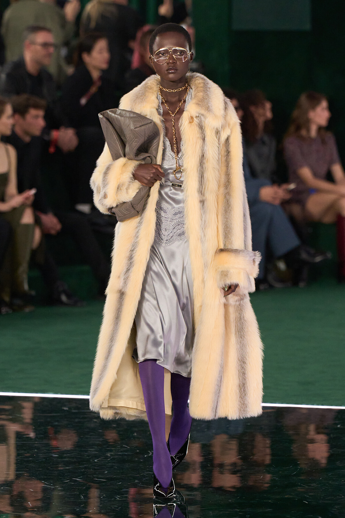 Runway model in a long cream fur coat over a silver dress, purple tights, bold jewelry, and oversized sunglasses, clutch under arm.
