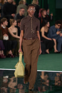 Model walking a runway in a brown top and tan wide-leg pants, accessorized with oversized glasses and a pale yellow handbag.