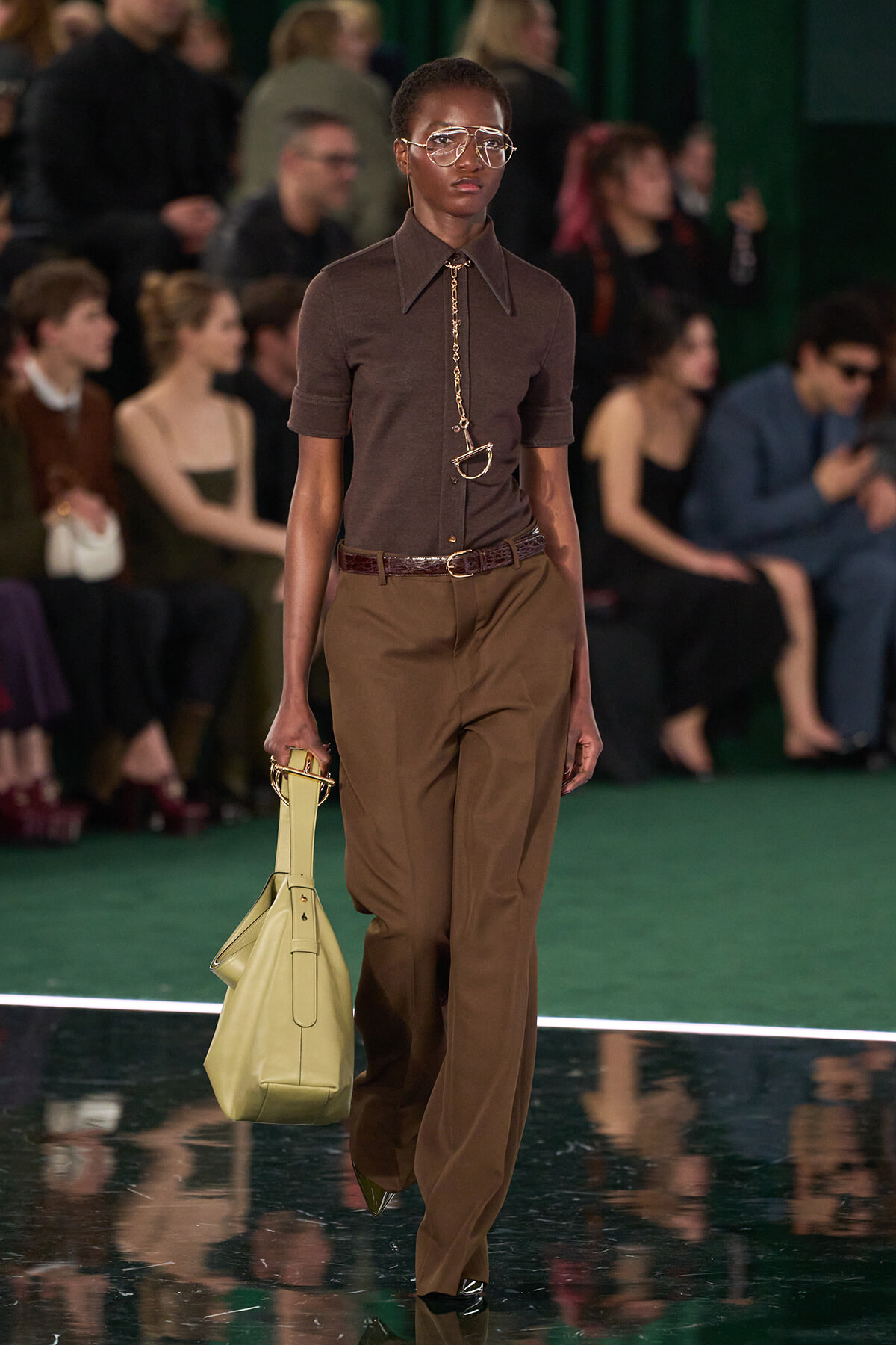 Model walking a runway in a brown top and tan wide-leg pants, accessorized with oversized glasses and a pale yellow handbag.