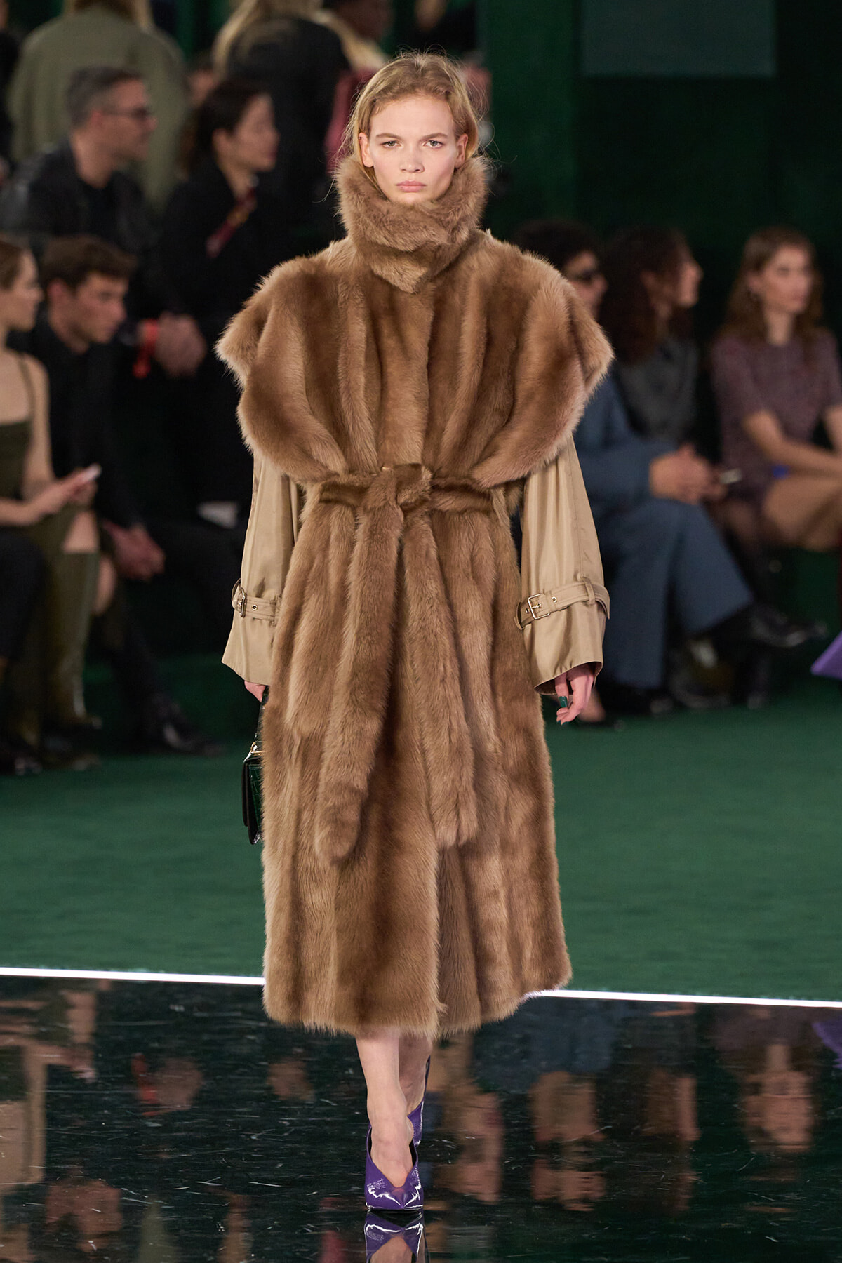 Model on a runway wearing a full-length brown fur coat with a belt; audience sits in the background.
