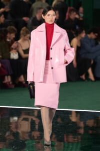 Model on a runway wearing a glossy pink oversized coat over a matching pink skirt, with a red turtleneck, clear glasses, and a black clutch.