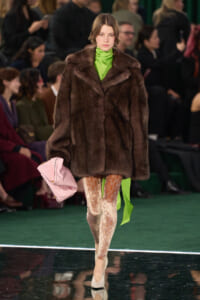 Model on a runway wearing a dark brown fur coat over a bright neon green turtleneck, lace tights, nude heels, and a pale pink handbag.