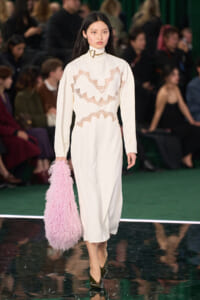 Model walks the runway in a cream turtleneck dress with pink lace panels, carrying a pink shaggy bag and black heels.