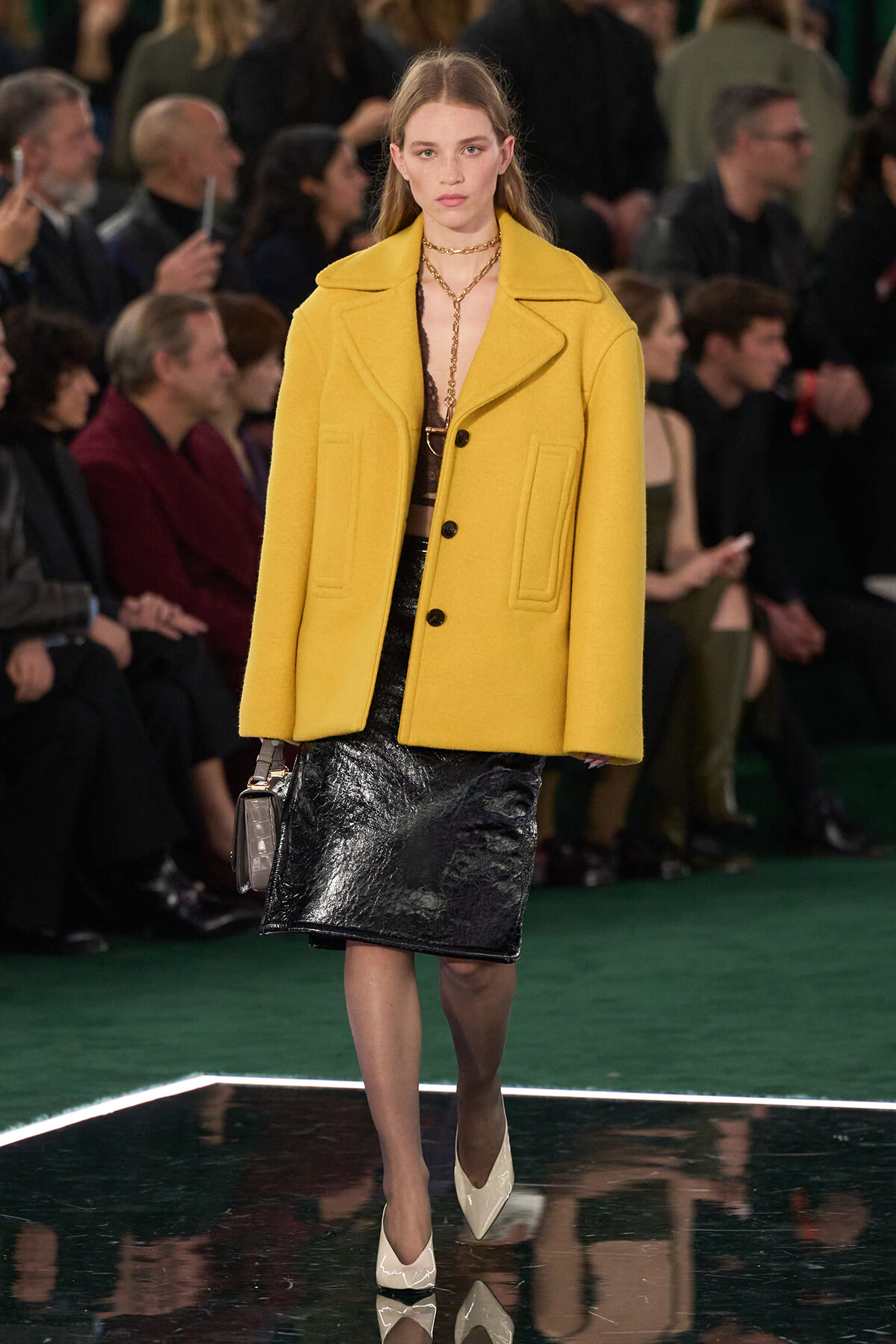Model walks the runway in a mustard-yellow oversized coat over a black shiny skirt, white heels, at a fashion show audience in the background.