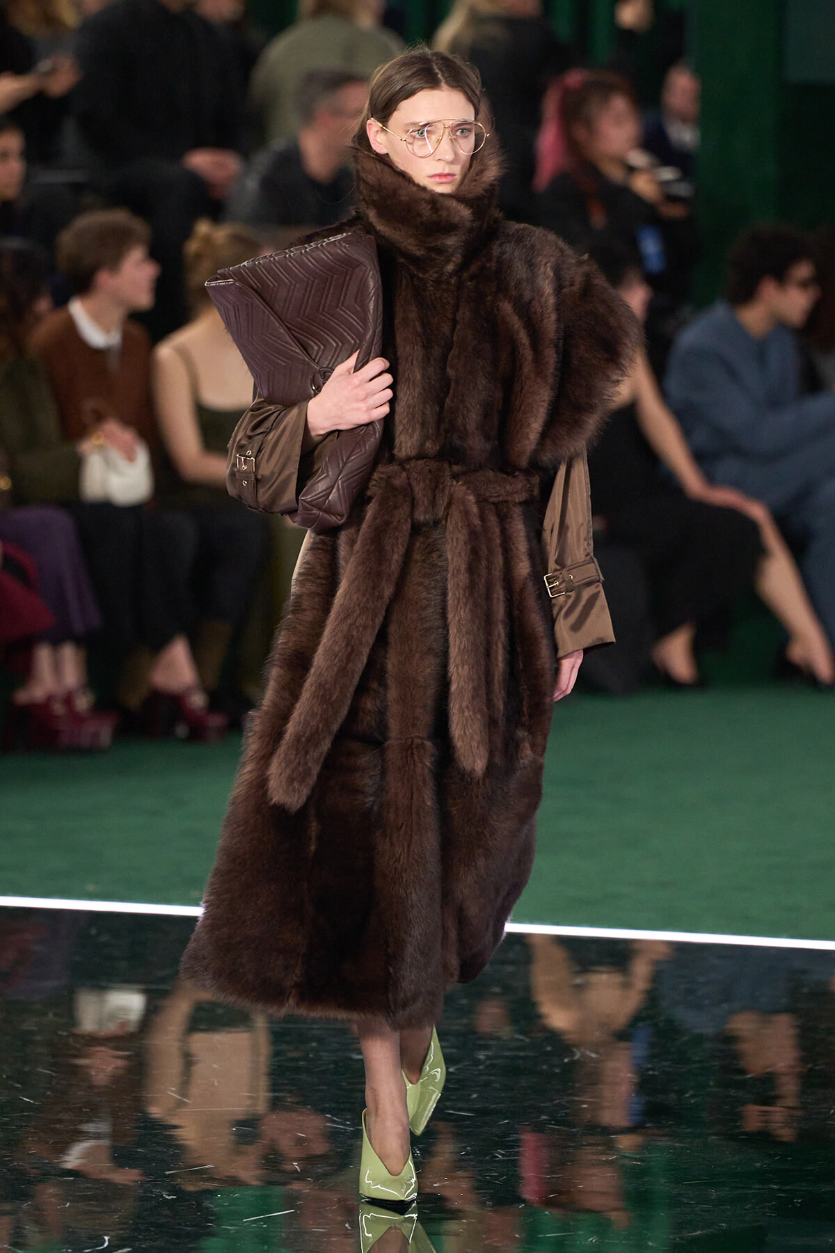 Model walking a runway in a dark brown fur coat with a large collar, gold-rimmed glasses, and a brown quilted clutch.