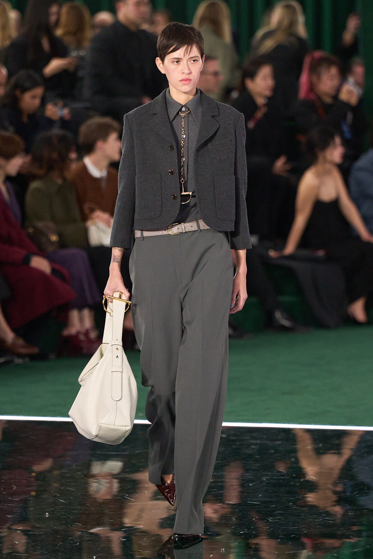 Model walks the runway in a charcoal cropped blazer, gray trousers, and a beige belt, carrying a white handbag.