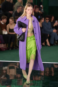 Model on a runway wearing a lavender coat over a lime green lace-trim dress, gold jewelry, pale green heels, and a black quilted clutch.