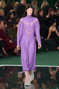 Model walks a runway wearing a lavender midi dress with a lace chevron panel and high neck; fluffy white shoes, audience in the background.