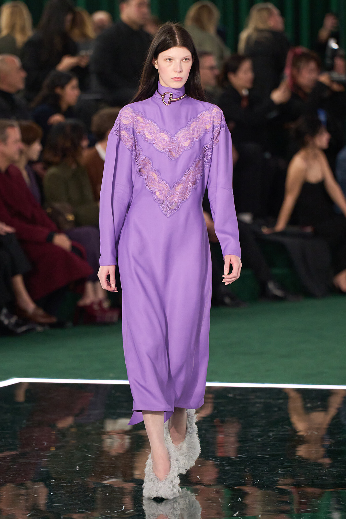 Model walks a runway wearing a lavender midi dress with a lace chevron panel and high neck; fluffy white shoes, audience in the background.