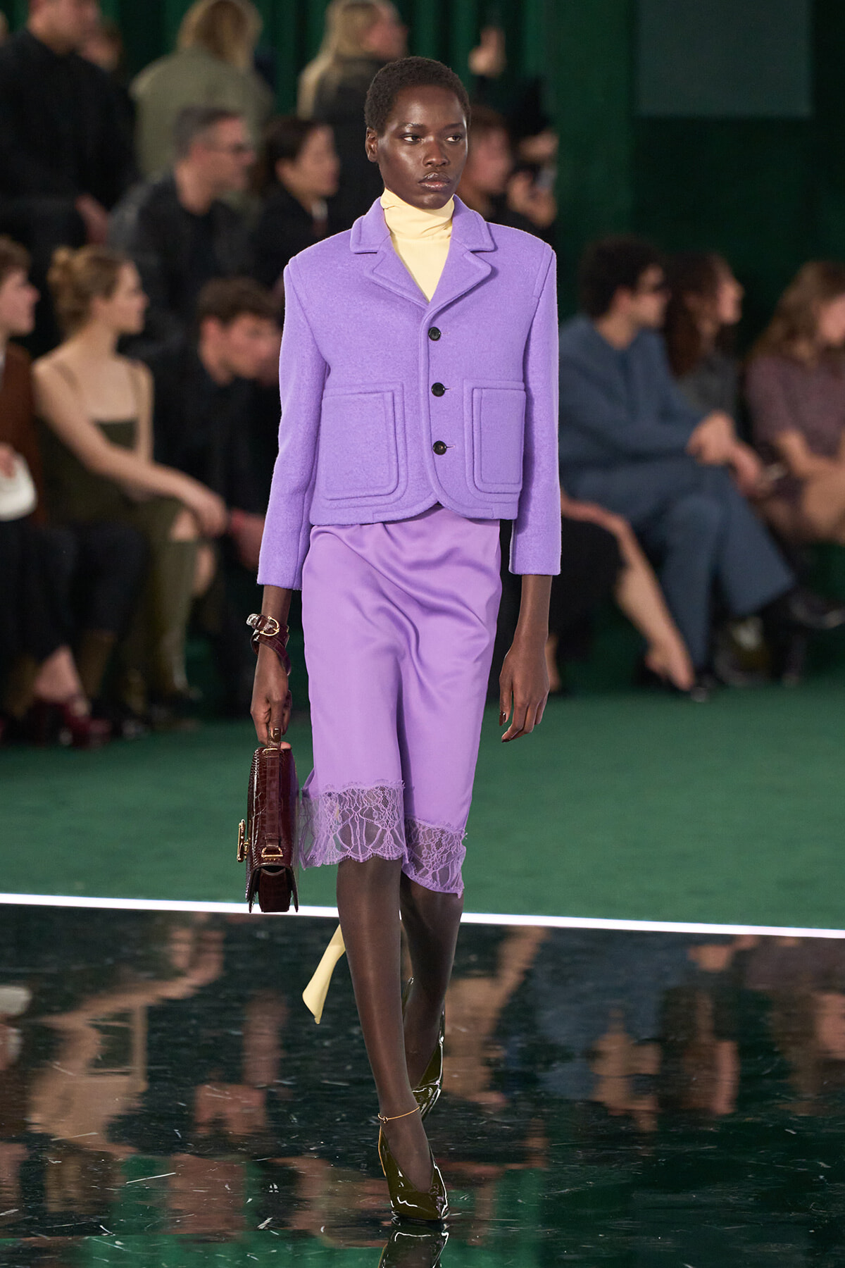Model walking a runway in a lavender suit with a matching jacket and knee-length lace-hem skirt, carrying a brown structured handbag.