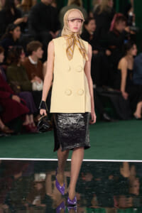 Model walking a fashion runway in a cream sleeveless coat with large gold ring details, a shiny black pencil skirt, beige headscarf, and purple pointed heels.