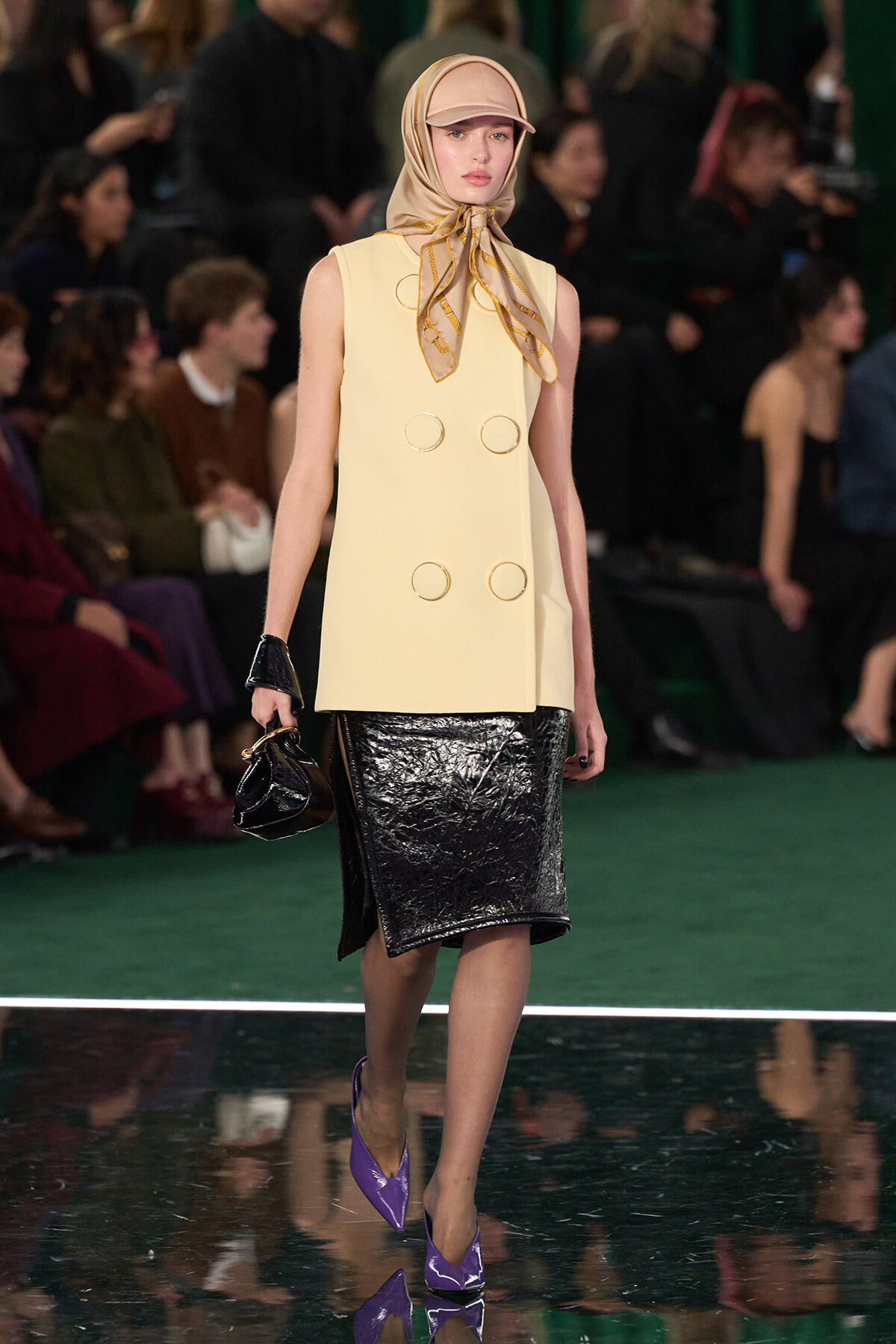 Model walking a fashion runway in a cream sleeveless coat with large gold ring details, a shiny black pencil skirt, beige headscarf, and purple pointed heels.