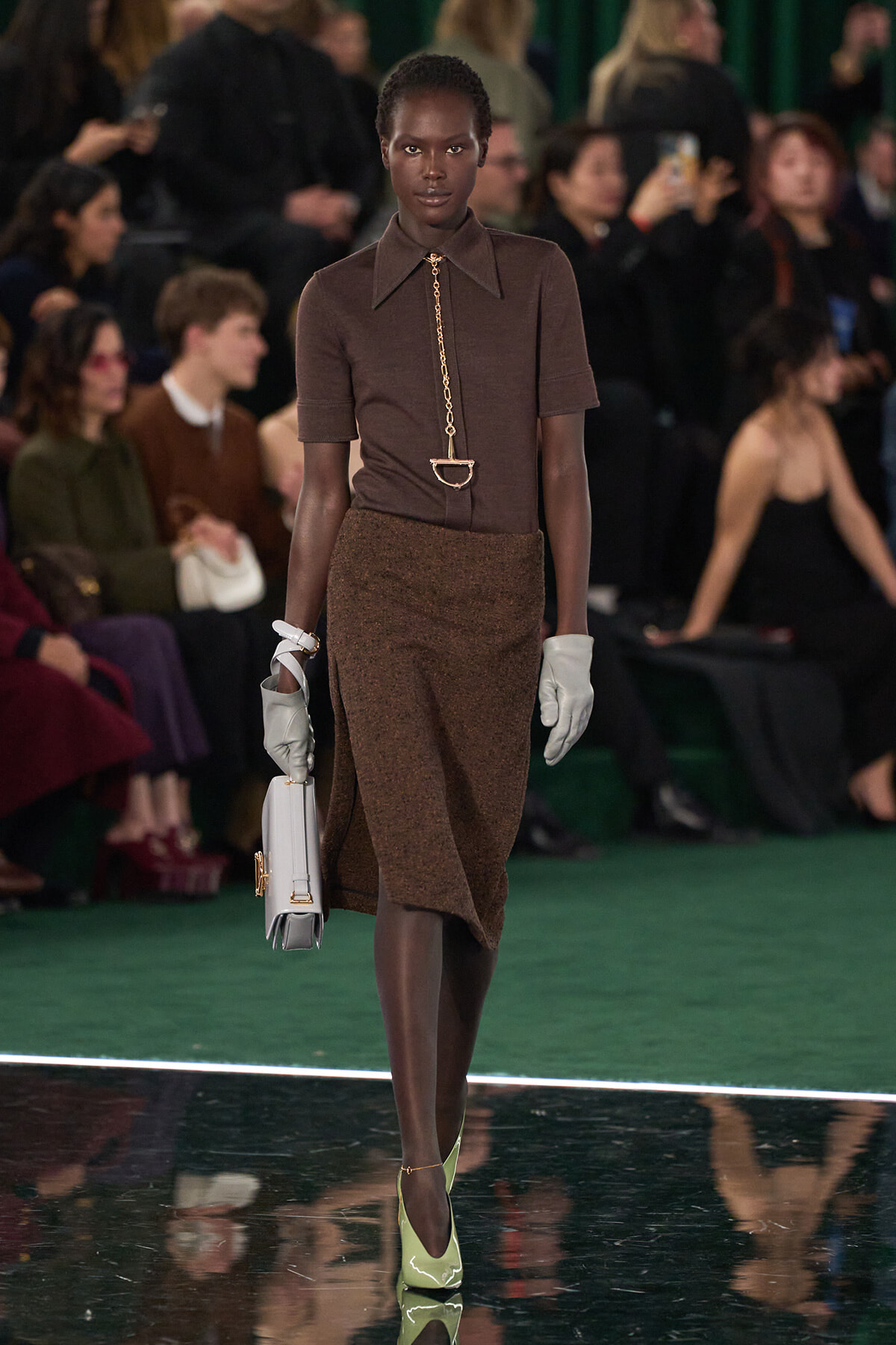 Black female model walks a runway in a brown short-sleeve top, textured brown skirt, white gloves, lime-green heels, and a light gray handbag.