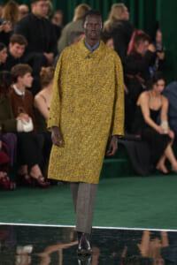 Male model walking a runway in a yellow textured coat, grey pants, and black boots; audience visible in background.