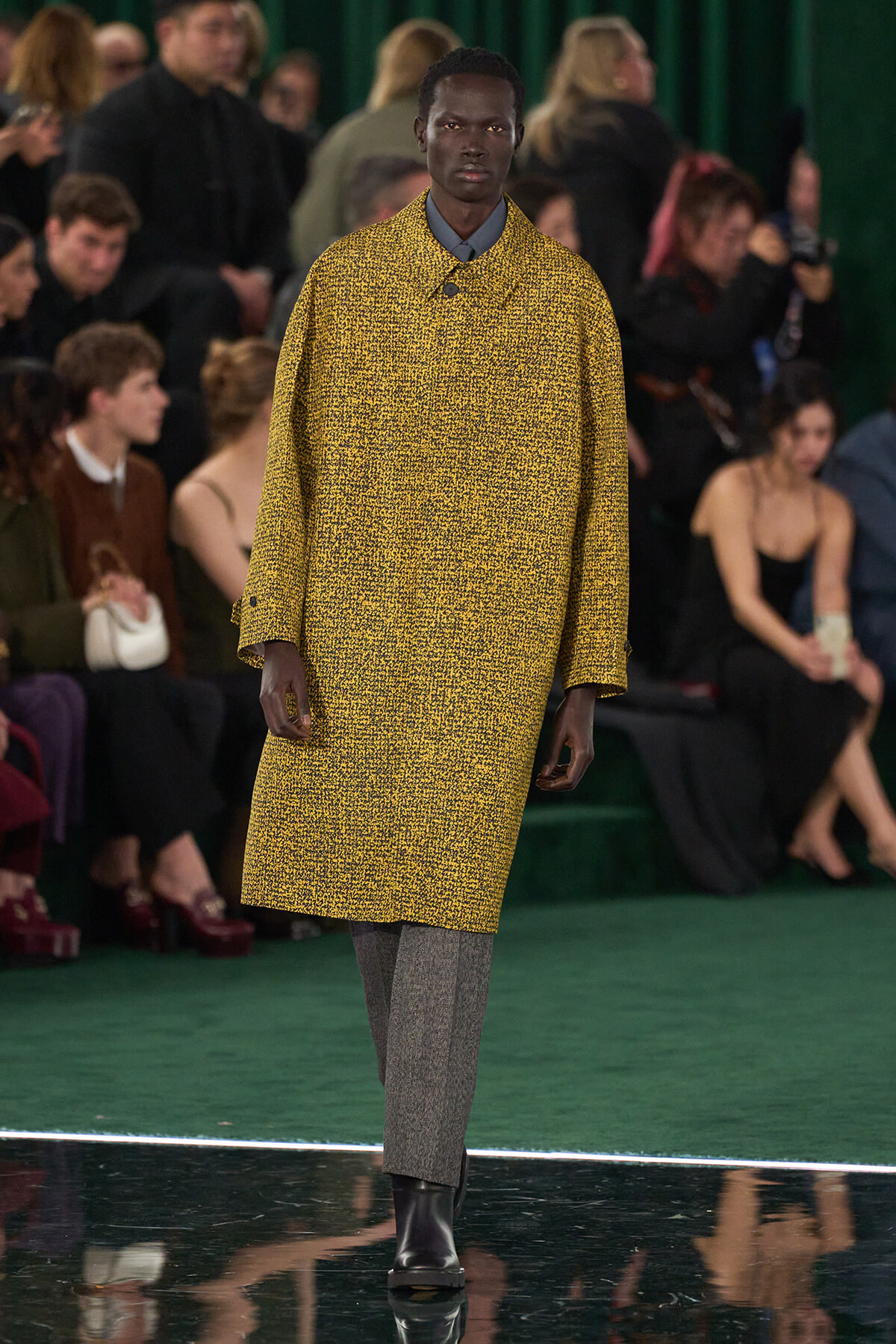 Male model walking a runway in a yellow textured coat, grey pants, and black boots; audience visible in background.