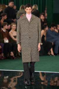 Male model walks a fashion show runway in a speckled beige overcoat and pale pink turtleneck, with blurred audience in the background.