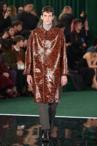 Male model walks the runway wearing a shiny leopard-print coat over a gray turtleneck, with an audience watching in the background.