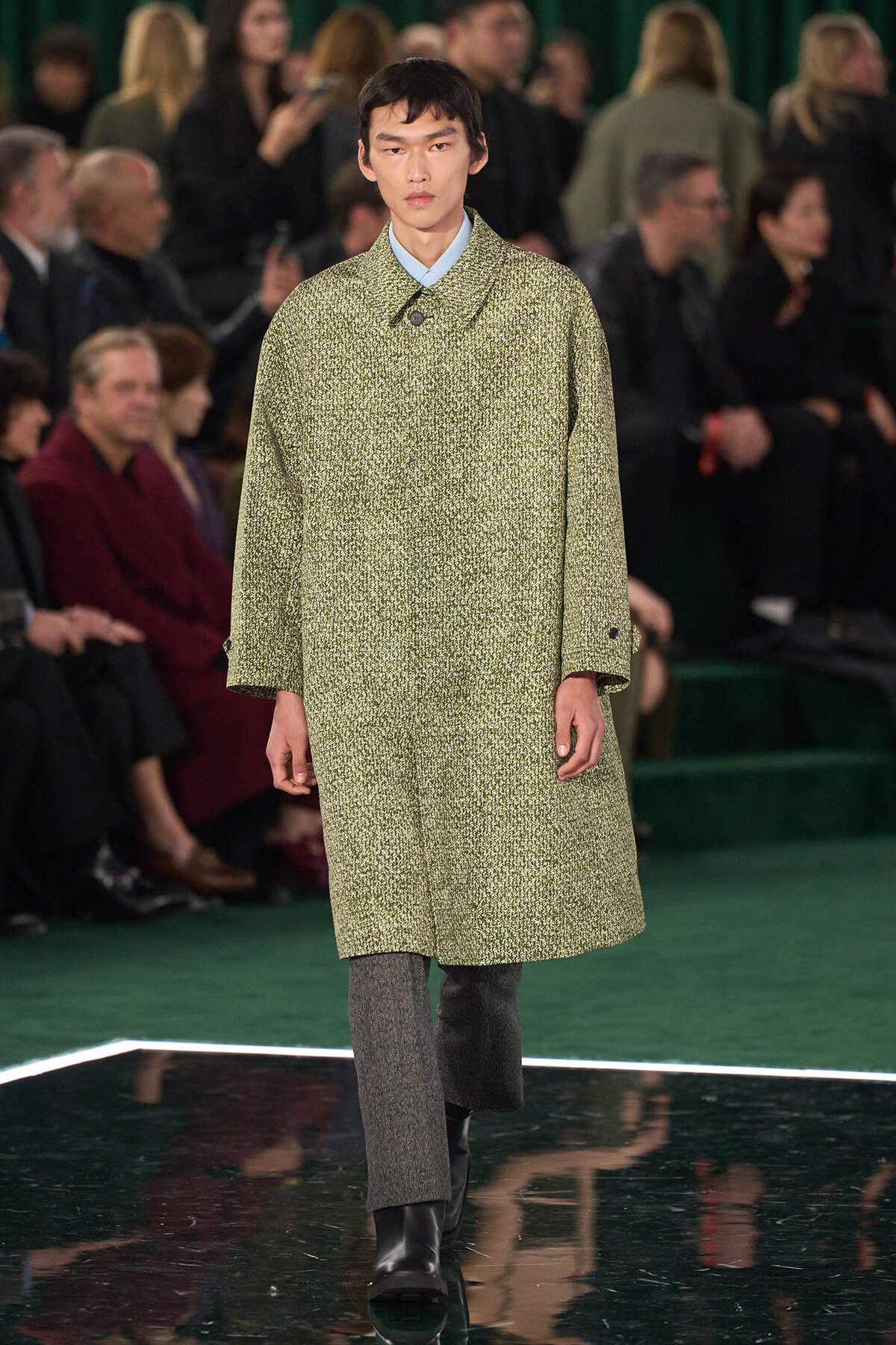 Male model walks a fashion show wearing a green speckled long coat, light blue shirt collar visible, gray pants, black boots in front of a seated audience.
