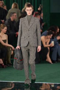 Male model on a fashion runway wearing a speckled grey suit with turtleneck, clear glasses, and carrying a large gray duffel bag; green runway in background.