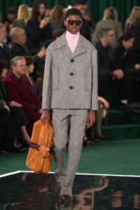 Male model on a fashion runway wearing a gray textured suit with a pink turtleneck, dark sunglasses, and a tan duffel bag.