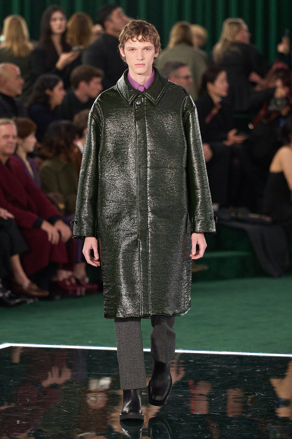 Male model walking a runway in a dark metallic overcoat, purple shirt, gray pants, black boots; audience in the background is blurred.