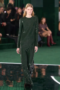 Model walking the runway in a glittery dark green full‑length jumpsuit on a fashion show stage.
