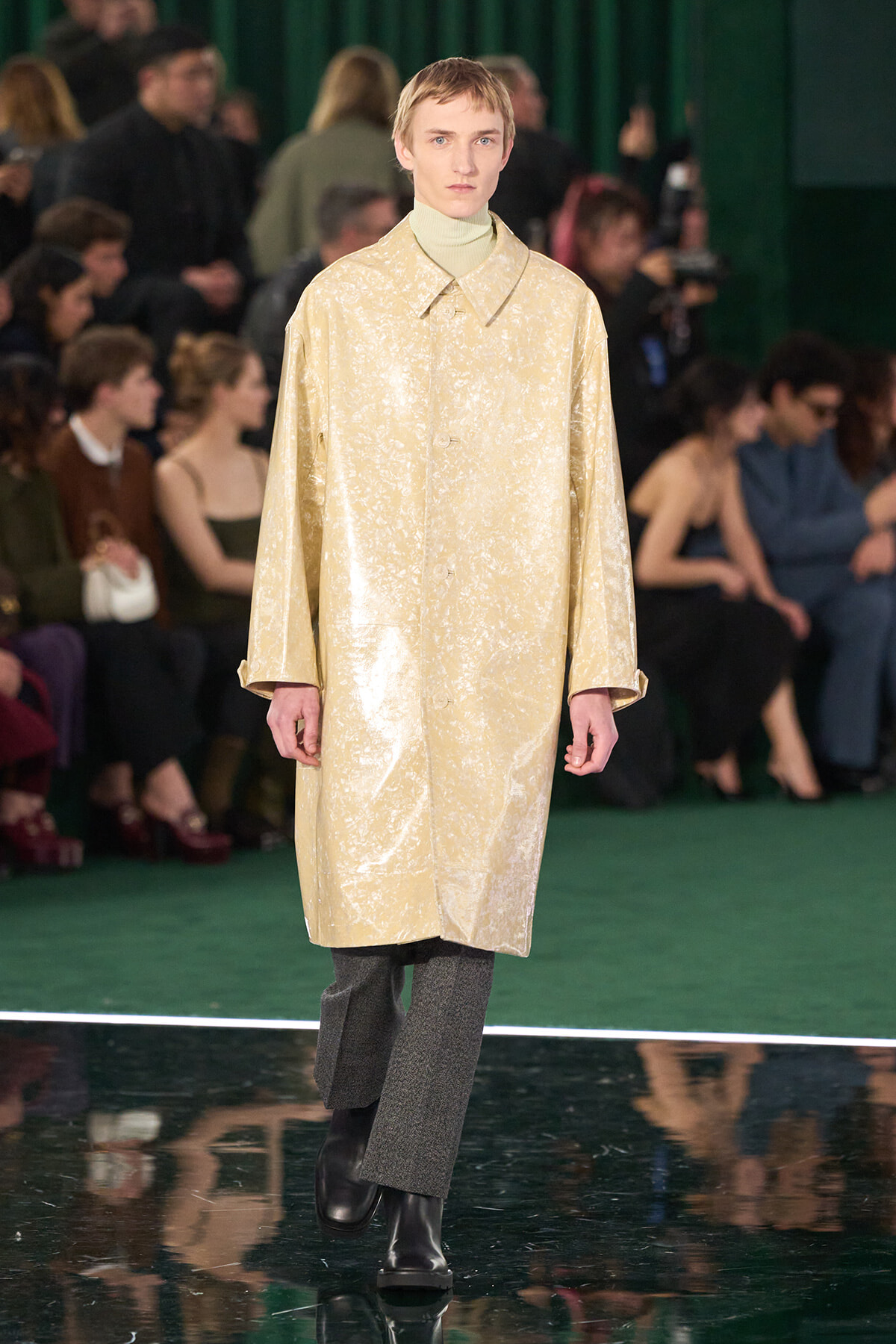 Model walking the runway in a shiny beige coat over a pale turtleneck, with spectators in the background.