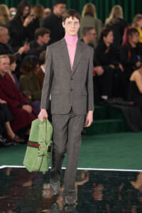 Male model in a gray suit with a pink turtleneck walks the runway, carrying a green duffel bag and wearing clear glasses in a fashion show setting.