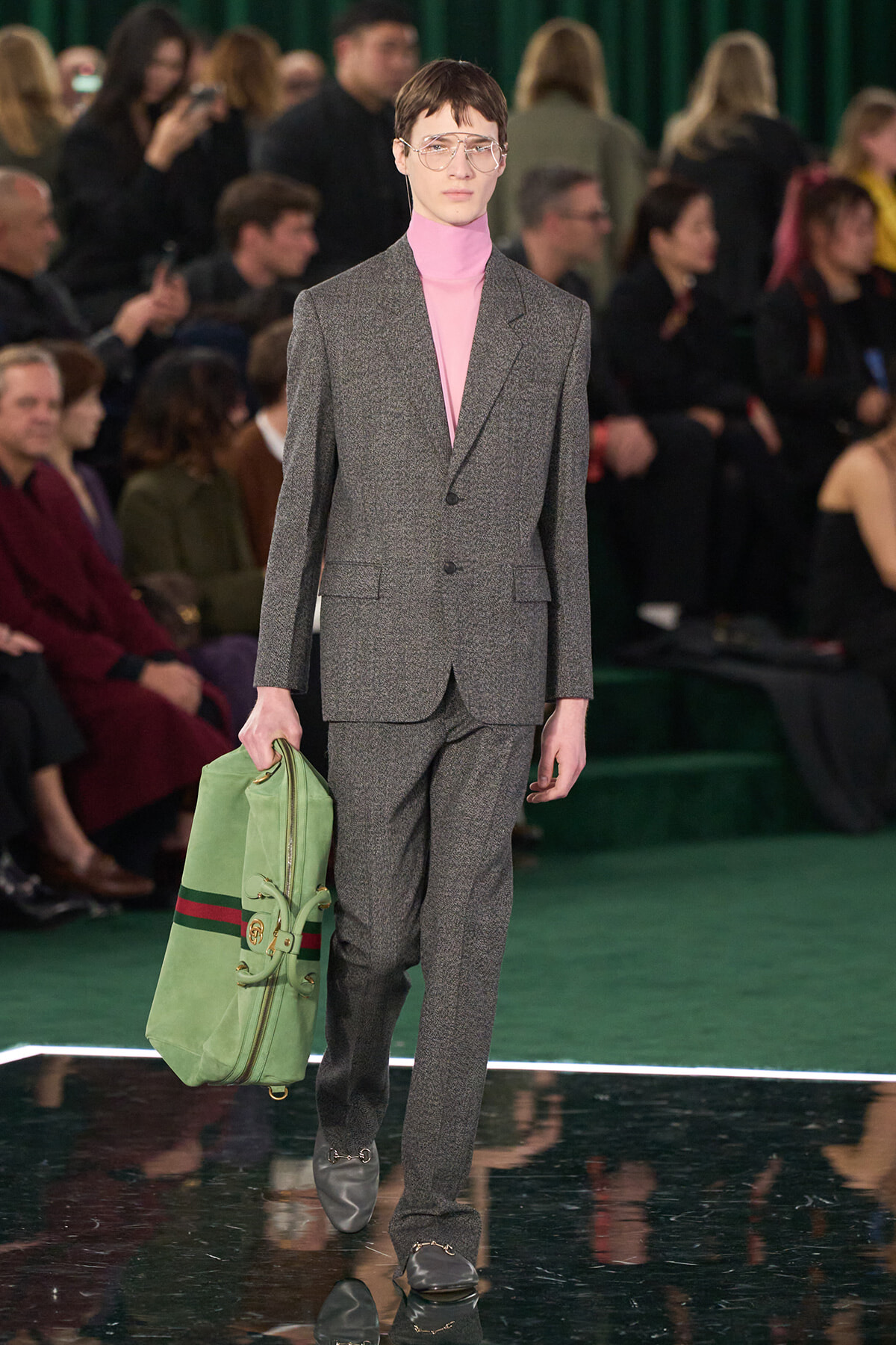 Male model in a gray suit with a pink turtleneck walks the runway, carrying a green duffel bag and wearing clear glasses in a fashion show setting.