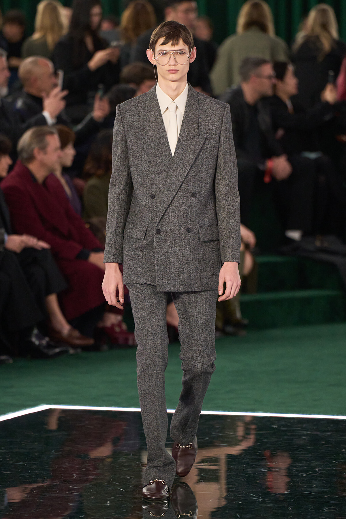 Male model walking the runway in a gray textured double‑breasted suit with a white shirt and clear glasses, audience in the background.