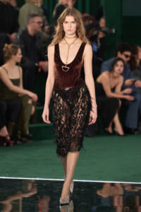 Model walking the runway in a burgundy velvet sleeveless top and black lace skirt with a gold chain necklace.