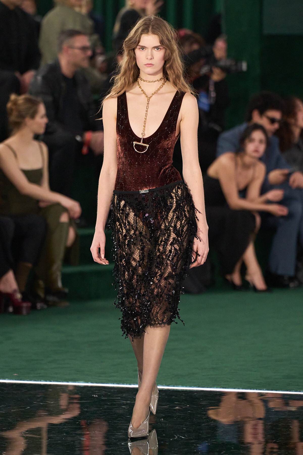 Model walking the runway in a burgundy velvet sleeveless top and black lace skirt with a gold chain necklace.
