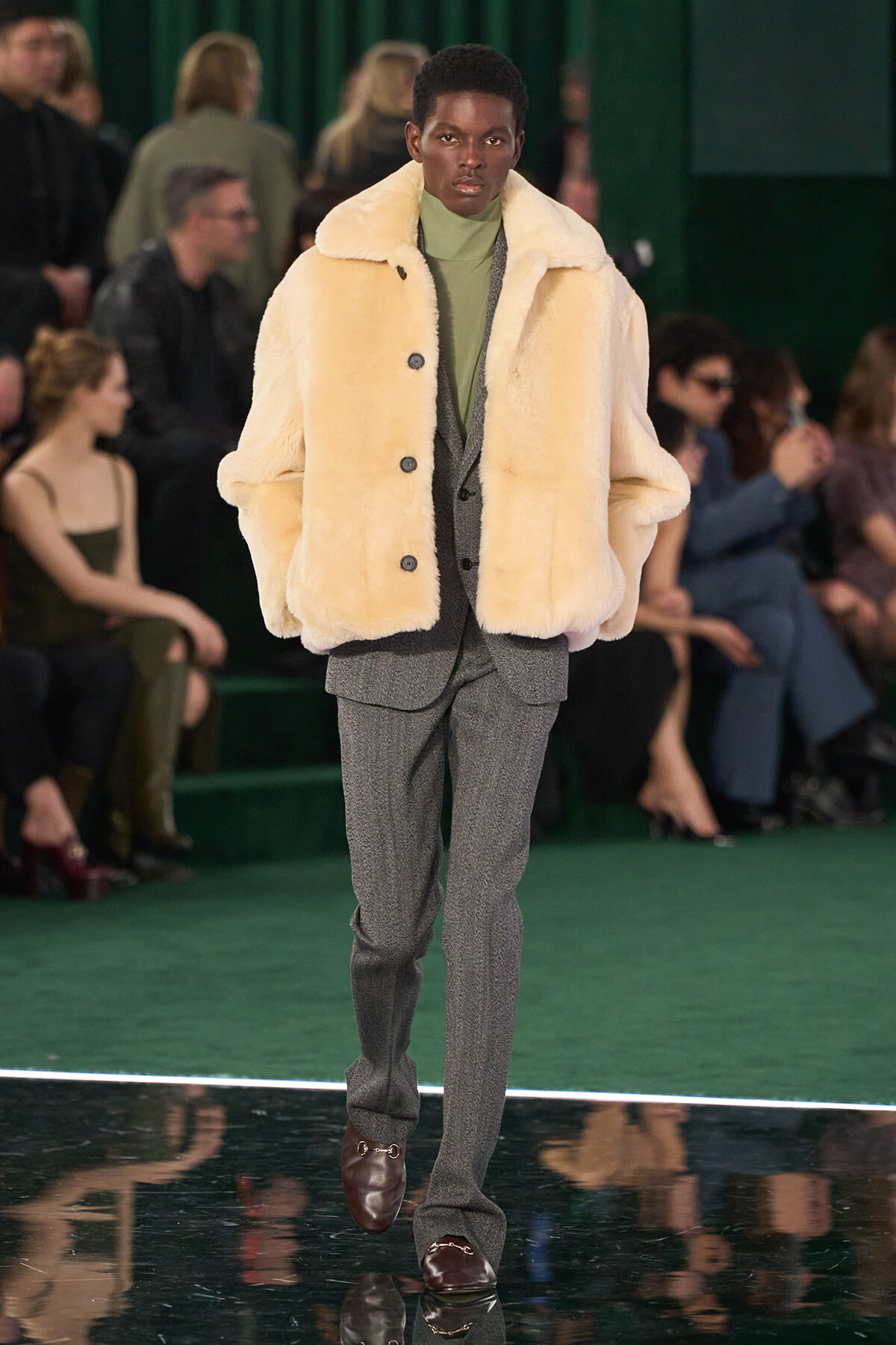 Male model walks the runway in a cream faux‑fur coat over a grey suit with a green turtleneck; audience blurred in the background.