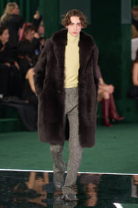Model walks a runway in a dark fur coat over a pale yellow turtleneck and glittery gray pants.