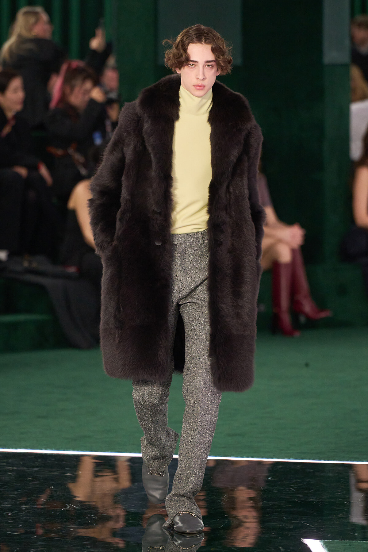 Model walks a runway in a dark fur coat over a pale yellow turtleneck and glittery gray pants.