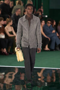 Male model on a fashion runway wearing a gray fuzzy cardigan, brown shirt and tie, dark trousers, green loafers, and a pale yellow bag.