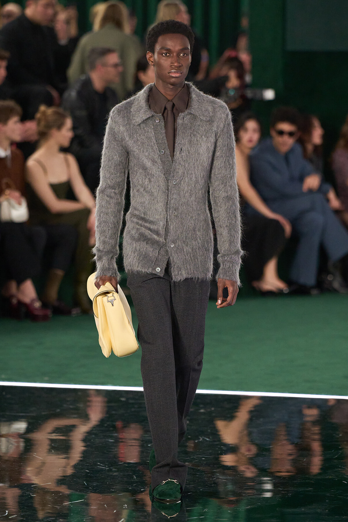 Male model on a fashion runway wearing a gray fuzzy cardigan, brown shirt and tie, dark trousers, green loafers, and a pale yellow bag.