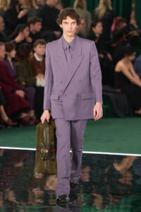 Male model in a lavender double-breasted suit walking a runway, carrying a green leather duffel bag; audience blurred in the background.