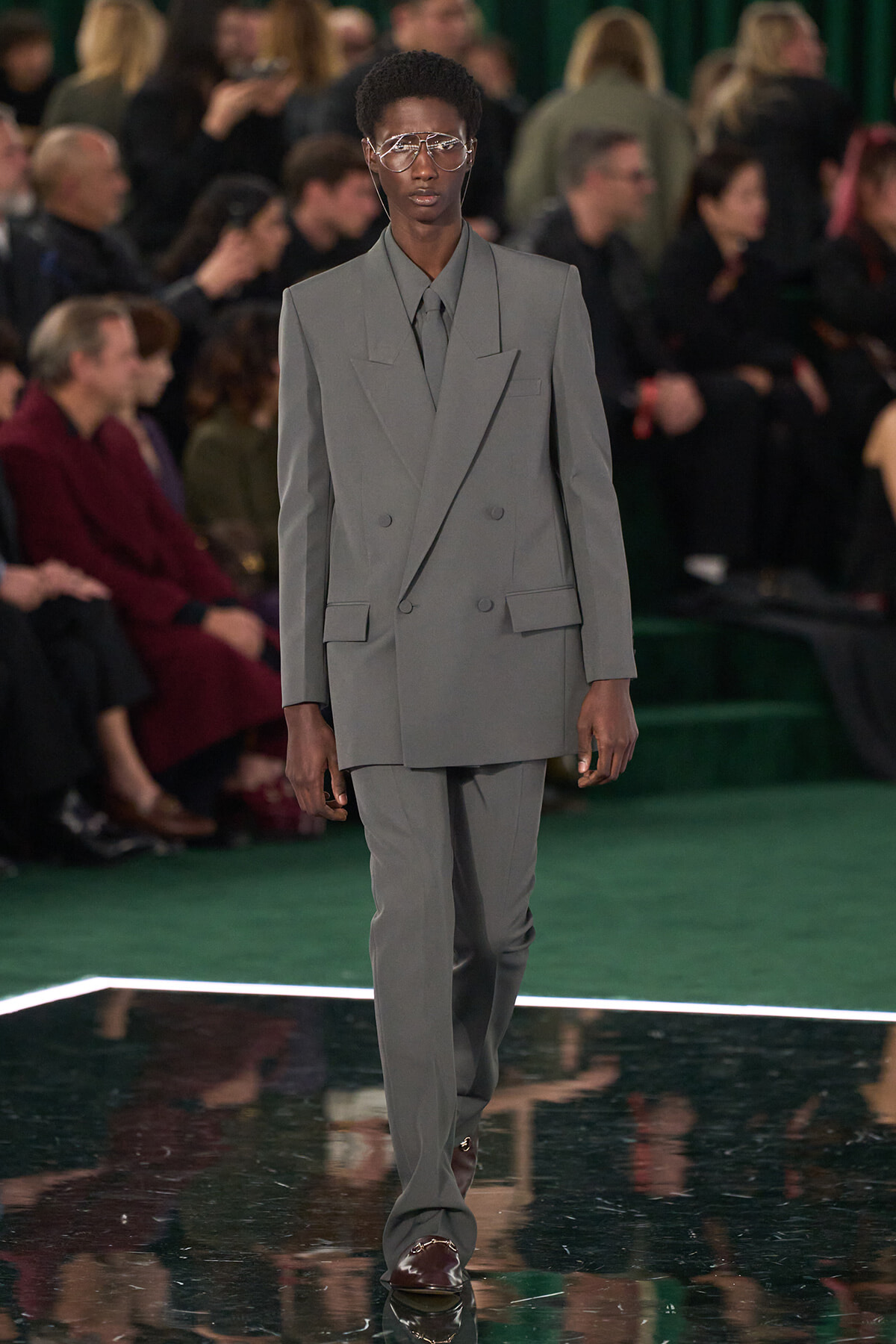 Model walks a runway in a light gray oversized double-breasted suit with wide lapels and clear-framed glasses; audience out of focus in background.