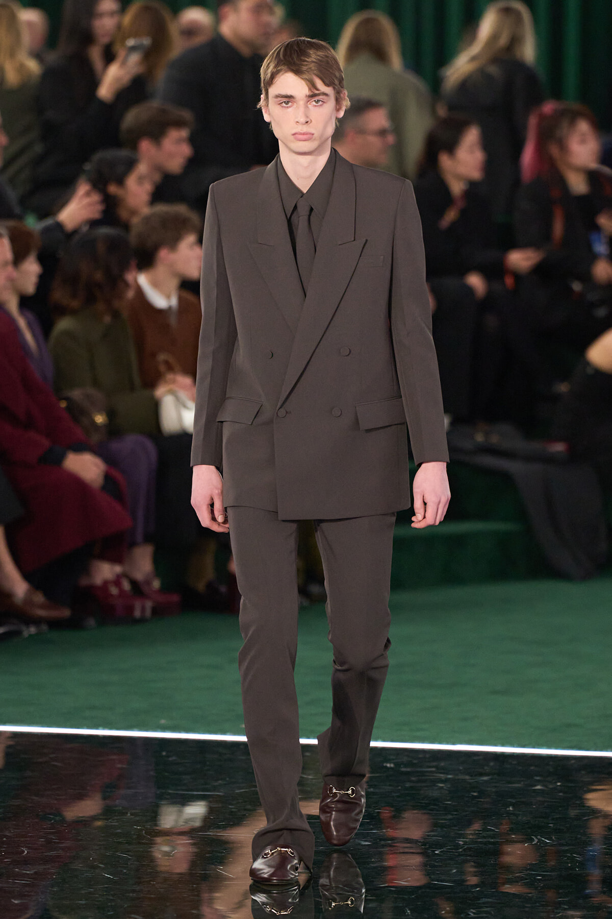 Male model walking a runway in a dark gray double-breasted suit with a black shirt and loafers, audience in the background
