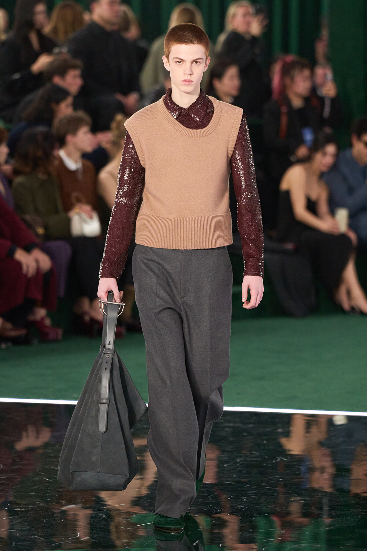 Male model walks a runway in a camel knit vest over a burgundy sequined shirt, gray trousers, and carries a large gray bag; audience in background