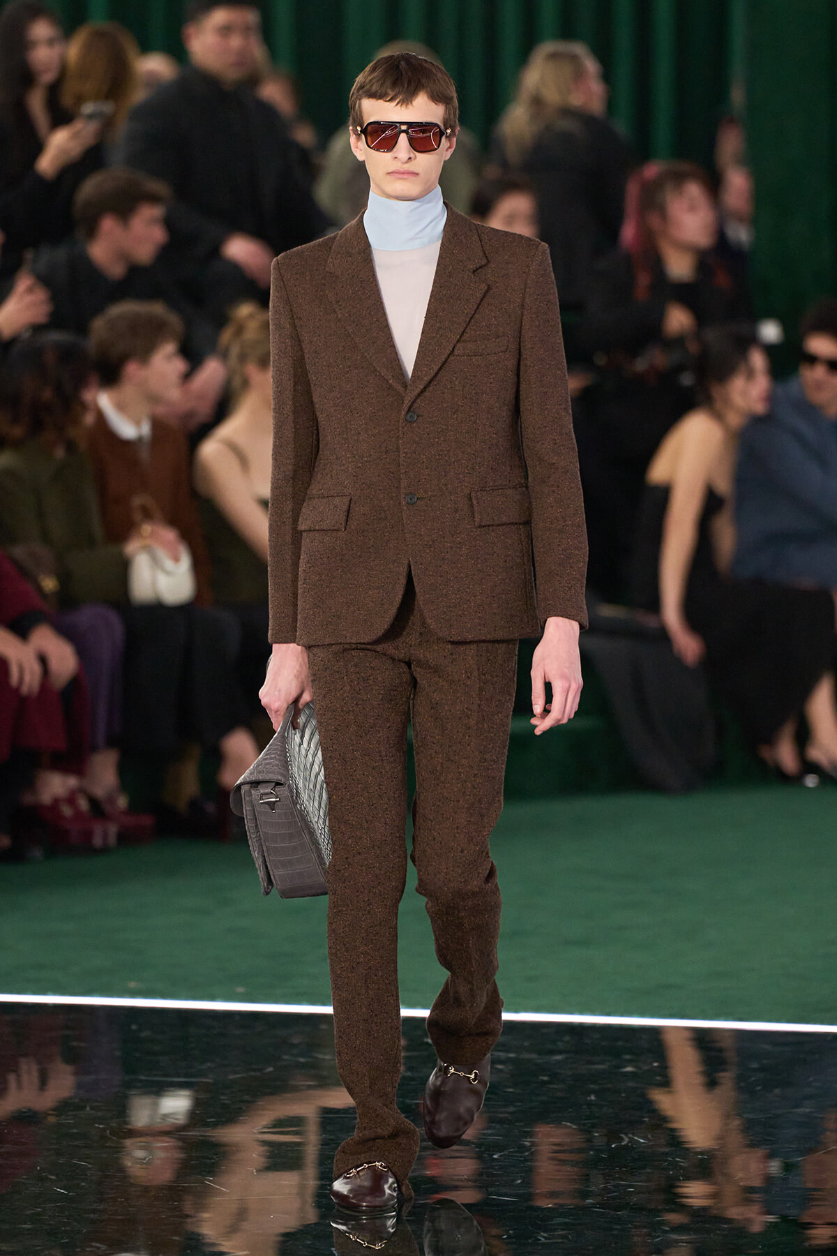 Model walking a fashion runway in a brown tailored suit with a light blue turtleneck, dark sunglasses, and a gray crocodile-embossed clutch.