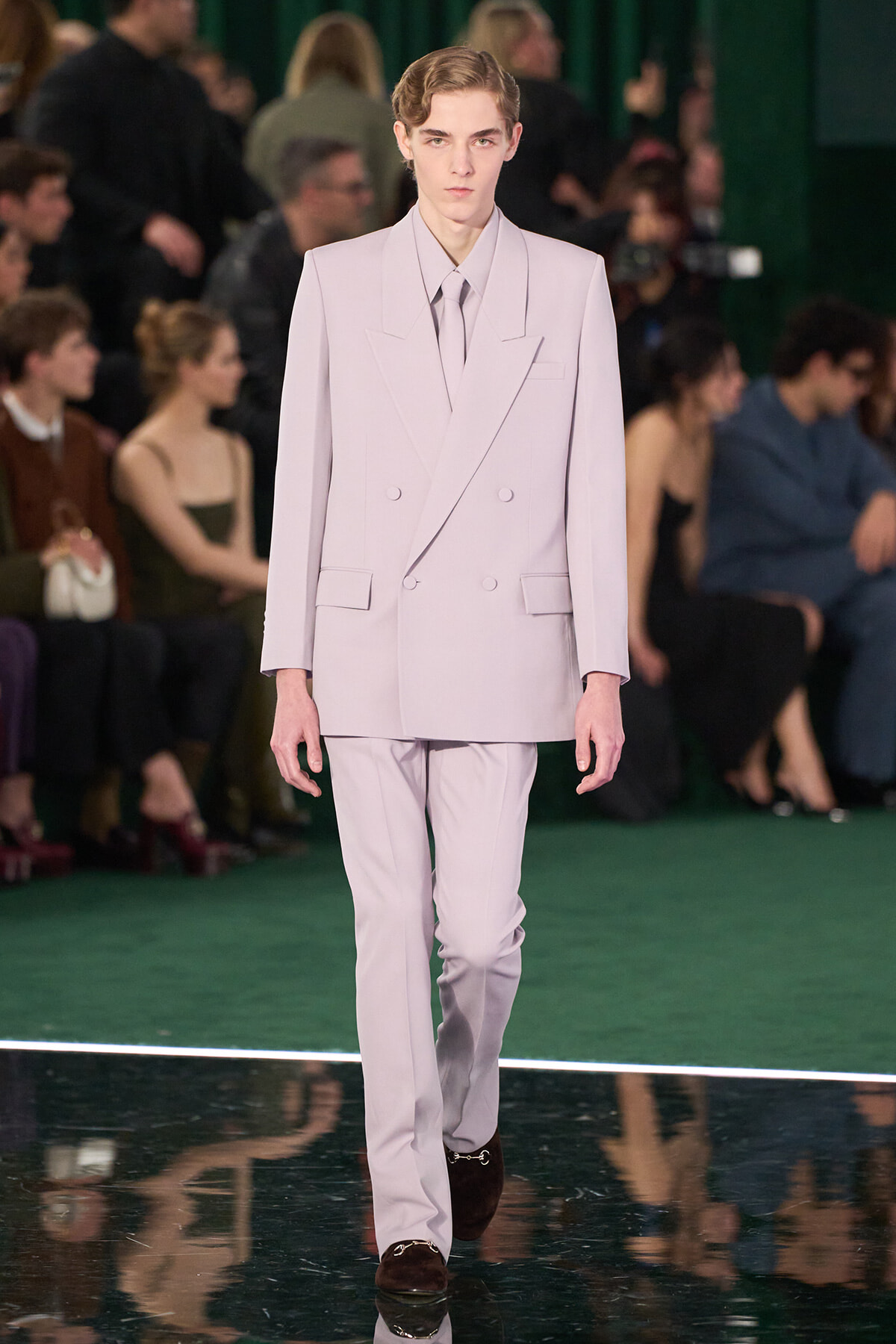 Male model walks the runway in a light lavender double-breasted suit and tie, on a green catwalk with spectators in the background.